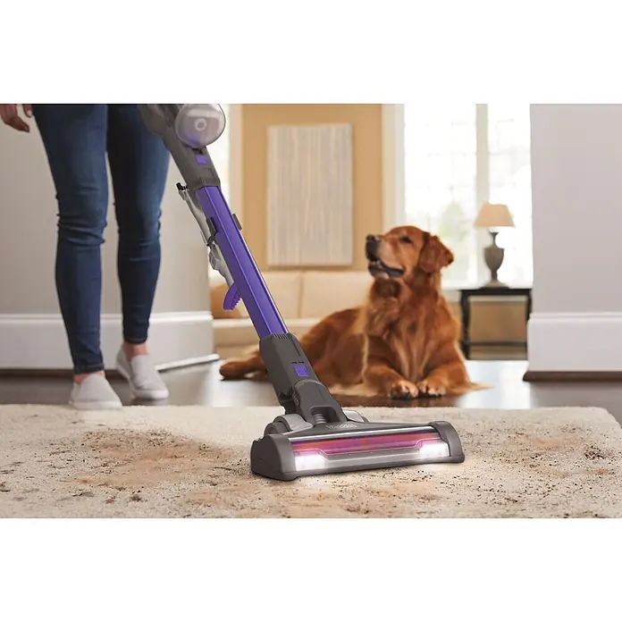 Black+Decker Aspirador de batería Power Series Extreme - 18 V, 2 Ah, Volumen de recipiente: 650 ml - Una persona aspira una alfombra sucia con una aspiradora de escoba morada y gris con cabezal iluminado. Un perro observa desde el fondo.