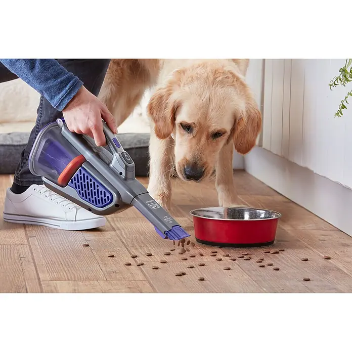 Black+Decker Aspirador de mano de batería GEN 11 - 18 V, 2 Ah, Volumen de recipiente: 700 ml - Una persona usa una aspiradora de mano gris para limpiar comida de perro derramada en un suelo de madera, junto a un golden retriever.