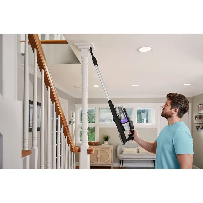 Black+Decker Aspirador de batería BSV520BRP-QW - 18 V, 1 batería, 2 Ah, Volumen de recipiente: 0,65 l - Hombre limpia una moldura alta con una aspiradora escoba inalámbrica negra y plateada con detalles morados y tubo de extensión.
