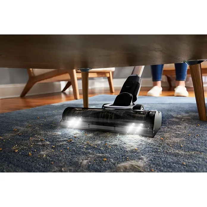Black+Decker Aspirador de batería BSV525BLP-QW - 18 V, 1 batería, 2,5 Ah, Volumen de recipiente: 0,65 l - Un cabezal de aspiradora con luces LED brillantes ilumina suciedad y pelos de mascotas en una alfombra azul mientras limpia bajo una mesa de madera.