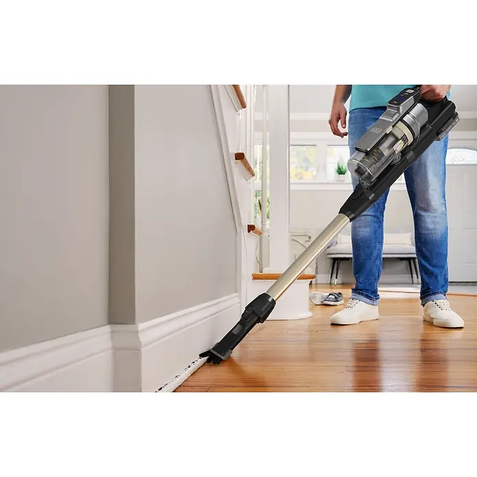 Black+Decker Aspirador de batería BSV525BLP-QW - 18 V, 1 batería, 2,5 Ah, Volumen de recipiente: 0,65 l - Persona usando una aspiradora escoba inalámbrica negra y plateada para limpiar polvo y suciedad de un suelo de madera junto a un zócalo.