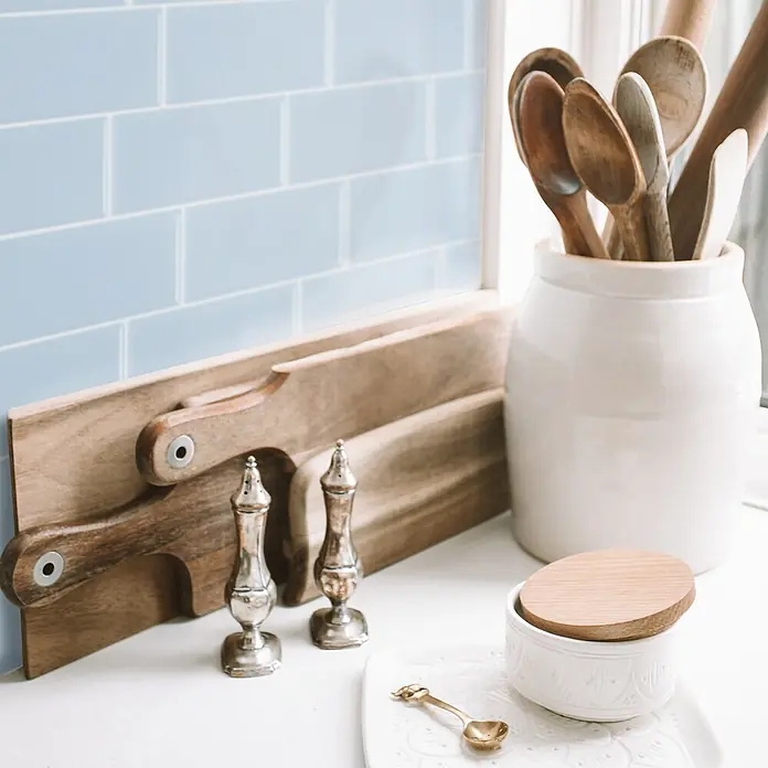 Smart Tiles Selbstklebemosaik Metro - 29,34 x 21,28 cm, Babe, Glänzend - Küchenarbeitsplatte mit hellblauen Metrofliesen, Holzbrettern, silbernen Streuern, Holzlöffeln im Topf und weißer Schale mit Deckel.