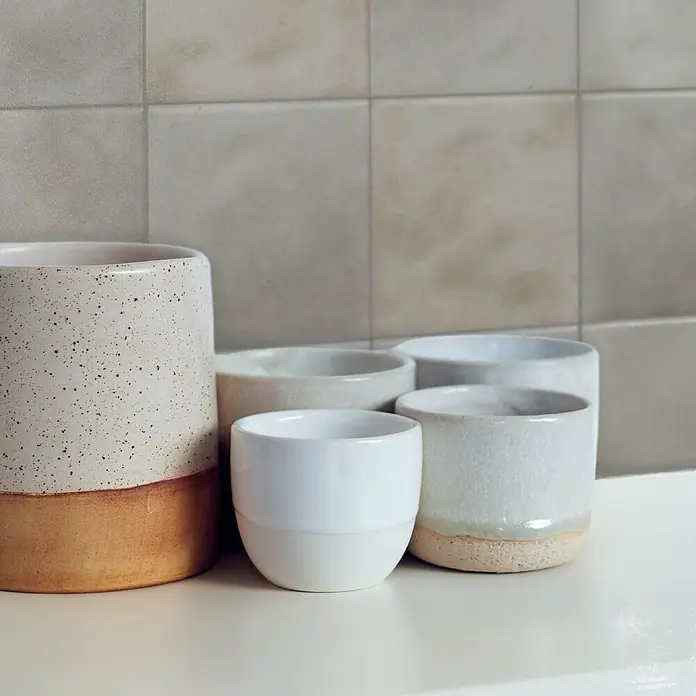 Smart Tiles Selbstklebemosaik Zellige - 22,86 x 22,86 cm, Fez, Matt - Eine Sammlung handgefertigter Keramikbecher und -töpfe in verschiedenen Größen und neutralen Tönen, teils gesprenkelt mit naturfarbenen Tonböden.