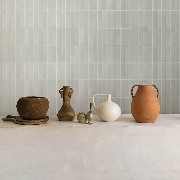 Smart Tiles Selbstklebemosaik Morocco - 29,02 x 22,86 cm, Essaouira, Glänzend - Eine Sammlung rustikaler, erdiger Keramikgefäße, darunter Krüge, Töpfe und ein Mörser mit Stößel, auf einem strukturierten Regal vor einer hellen Fliesenwand.