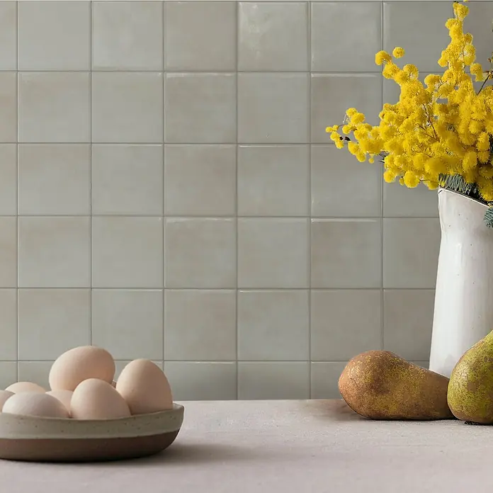Smart Tiles Selbstklebemosaik Zellige - 22,86 x 22,86 cm, Zagora, Matt - Hellgraue quadratische Fliesen als Küchenrückwand, mit Eiern und Obst auf der Arbeitsplatte.
