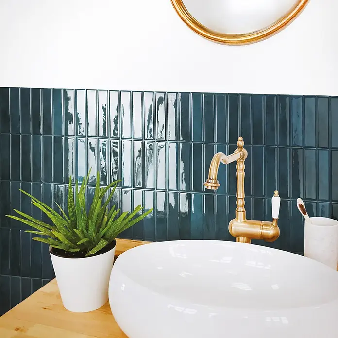 Smart Tiles Selbstklebemosaik Morocco - 29,02 x 22,86 cm, Agadir, Glänzend - Ein stilvoller Waschtisch mit einem weißen Aufsatzwaschbecken, einer goldenen Vintage-Armatur und glänzenden türkisfarbenen vertikalen Metro-Fliesen.