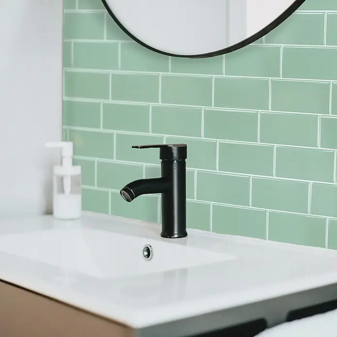 Smart Tiles Selbstklebemosaik Metro - 29,34 x 21,28 cm, Lucas, Glänzend - Mattschwarzer Wasserhahn über einem weißen Waschbecken vor einer Badezimmerwand mit glänzenden grünen Metrofliesen.