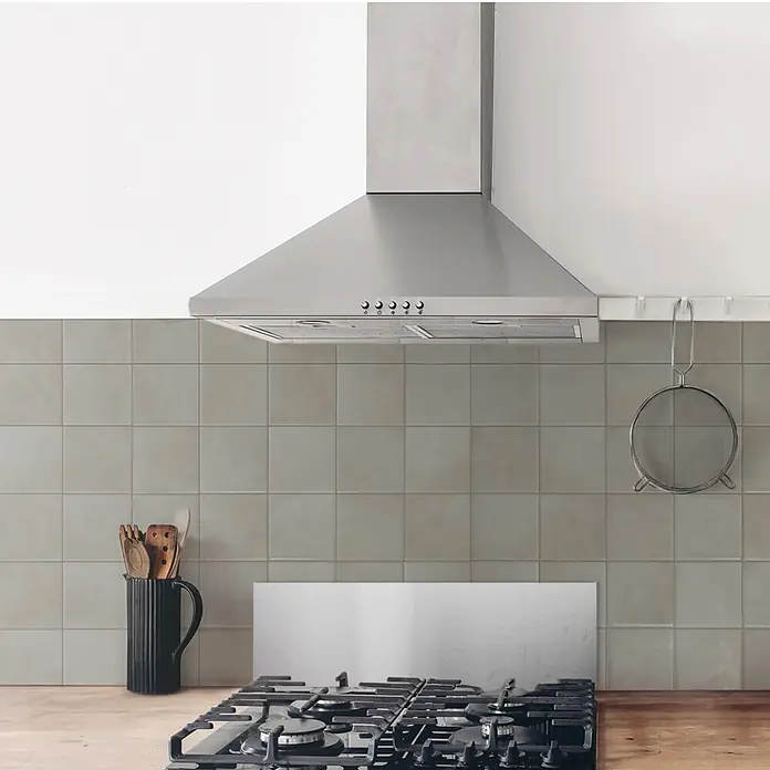 Smart Tiles Selbstklebemosaik Zellige - 22,86 x 22,86 cm, Zagora, Matt - Helle graue quadratische Küchenrückwandfliesen über einem Gasherd unter einer Dunstabzugshaube aus Edelstahl.