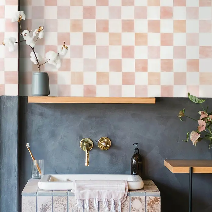 Smart Tiles Selbstklebemosaik Damier - 19,69 x 19,69 cm, Petra, Matt - Rosa-weiße Schachbrett-Wandfliesen über einem modernen Waschtischbereich mit Messingarmatur und Holzregal.