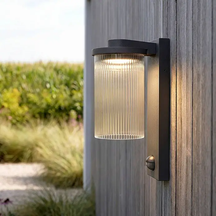 Lucande LED Wandleuchte aussen in schwarz, mit Bewegungsmelder, Aussenlampe -  - Dunkelgraue Außenwandleuchte mit geripptem Glasdiffusor und Bewegungssensor, an Holzzaun montiert.