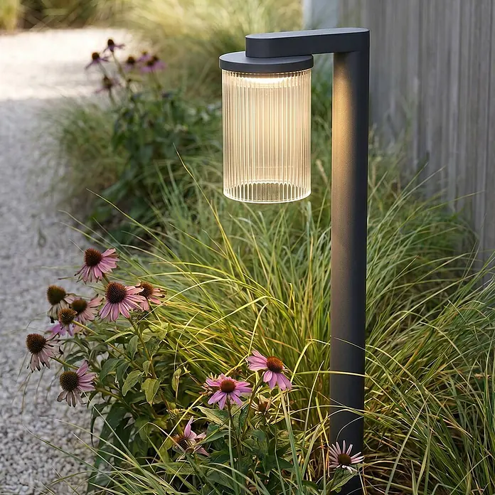 Lucande Außenlaterne Beelia -  - Dunkelgraue Außenleuchte mit geripptem Zylinderschirm, die warmes Licht abstrahlt, auf einem Gartenweg zwischen Pflanzen.