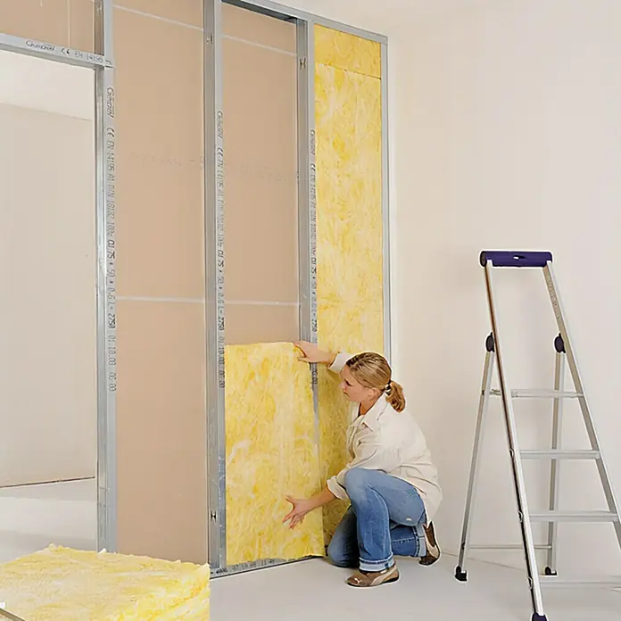 Probau Glaswolle Trennwandplatte TWP1 - Stärke: 100 mm, Wärmeleitfähigkeit: 0,039 W/(mK) - Eine Frau baut eine Trockenbauwand mit Dämmwolle und Gipskartonplatten, während eine Leiter im Hintergrund steht.