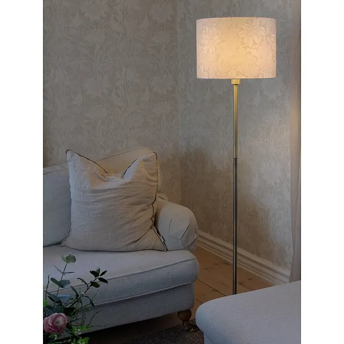 PR Home Stehleuchte -  - Elegante Stehlampe aus Messing mit dezent gemustertem hellen Schirm, die ein gemütliches Wohnzimmer neben einem Sofa beleuchtet.