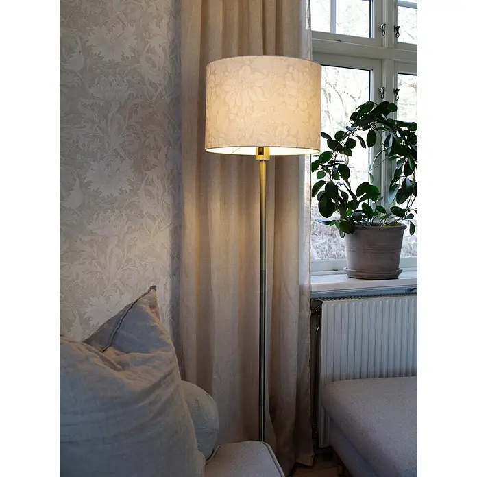 PR Home Stehleuchte -  - Eine goldene Stehlampe mit hellem, gemustertem Stoffschirm beleuchtet eine gemütliche Zimmerecke neben einem Fenster mit Vorhängen und einer Pflanze.