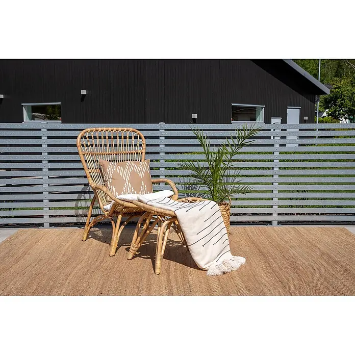 Ebuy24 Gartensessel Ella - 1 Stk., Rattan, 70% Polyester, 30% Baumwolle, B x T x H: 70 x 85 x 98 cm - Ein hellbrauner Rattan-Sessel mit Kissen und einer weißen Decke steht auf einem Jute-Teppich auf einer Terrasse.