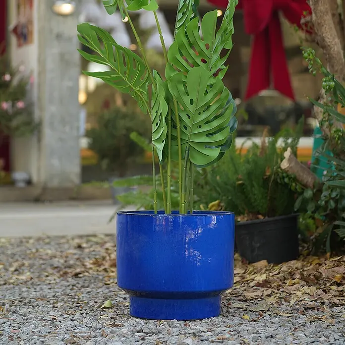 SONNENH Pflanztopf - Keramik, Blau - Ein leuchtend blauer Keramikpflanztopf mit einer künstlichen Monstera-Pflanze steht draußen auf Kies. Der Topf ist rund mit einem gestuften Sockel.
