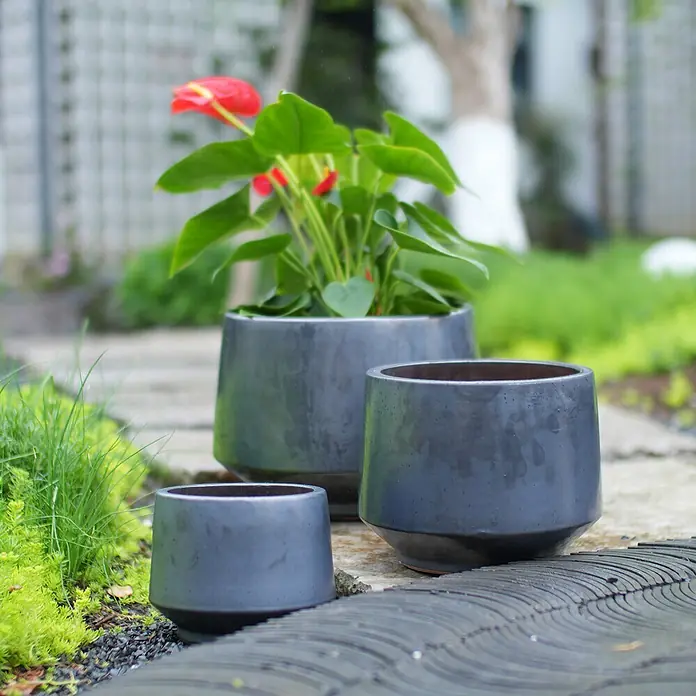 SONNENH Pflanztopf - 3 Stk., Keramik, Schwarz - Drei dunkelgraue Pflanzgefäße in verschiedenen Größen, eines mit einer rotblühenden Pflanze, auf einem Gartenweg.