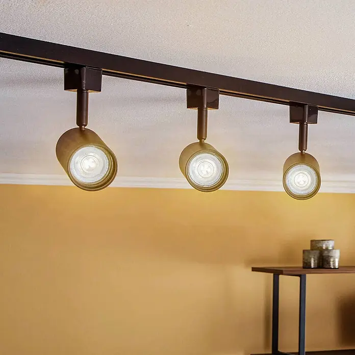 Prios LED-Spannseilsystem Jorell - schwarz, schwarz - Drei bronzefarbene Strahler an einem dunklen Schienensystem an der Decke, die eine warmgelbe Wand beleuchten.