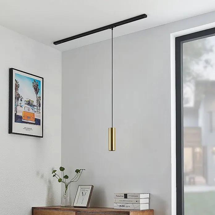 Lindby LED-Spannseilsystem Linaro - schwarz, messing / gold - Moderne goldene zylindrische Pendelleuchte hängt von einer schwarzen Deckenschiene über einer Holzkommode in einem Raum.