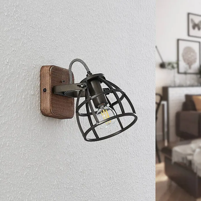 Lindby Wandleuchte innen in schwarz, braun, Wandstrahler -  - Industrielle Wandleuchte mit Holzsockel und dunklem Metallkäfig, mit sichtbarer Glühbirne, an einer Wand montiert.