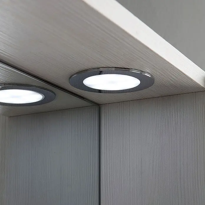 Kiamami Valentina LED-Spiegelschrank - B x H: 155 x 570 cm, Weiß - Nahaufnahme einer eingelassenen LED-Leuchte mit Chromblende, die helles weißes Licht unter einem hellen Holzregal abstrahlt, mit ihrer Reflexion in einem Spiegel.