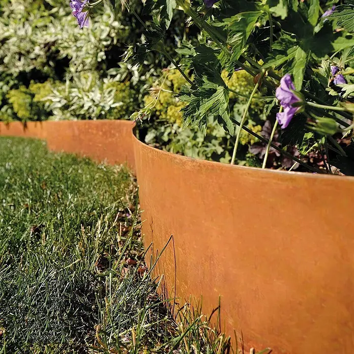 Bellissa Rasenkante Smoothedge - 500 x 15 cm, Rost, Metall - Gebogene Cortenstahl-Beeteinfassung trennt grünen Rasen von einem Blumenbeet mit violetten Blüten.