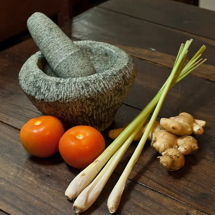 Deine Gartenwelt Zitronengras -  - Grauer Mörser und Stößel aus Stein mit Tomaten, Zitronengras und Ingwer auf Holz, bereit zum Zerkleinern von frischen Zutaten.