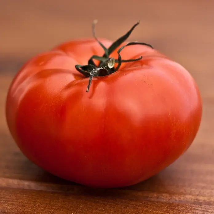 Deine Gartenwelt Gemüsesamen Fleischtomate - Solanum lycopersicum, Erntezeit: Juli - Eine frische, leuchtend rote Tomate mit grünem Stiel liegt auf einer Holzoberfläche.