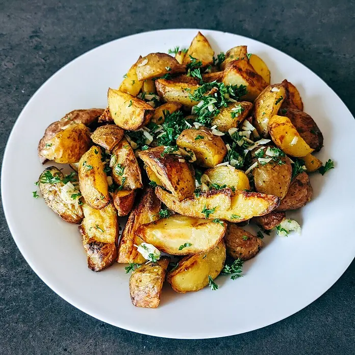 Deine Gartenwelt Petersilie Mooskrause 2 -  - Goldbraun geröstete Kartoffelspalten, garniert mit frischer Petersilie und gehacktem Knoblauch, auf einem weißen Teller.