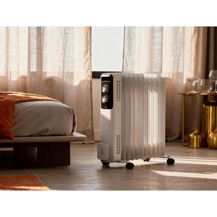 Purline Radiador de aceite Soleil - 1.000 W - 2.500 W, Blanco, 24 x 47 x 55 cm - Radiador de aceite blanco con perillas y ruedas, en el suelo de un dormitorio soleado junto a una cama.