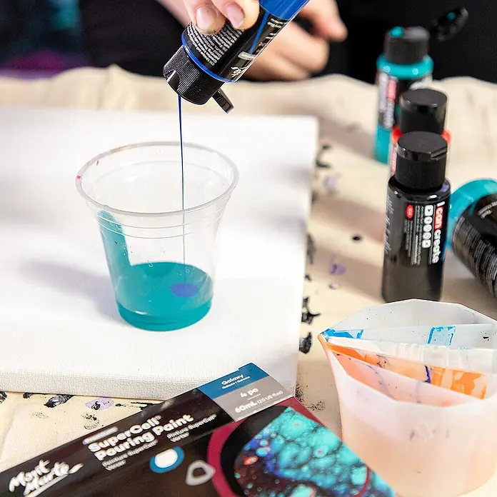 Mont Marte Acrylfarben-Set SuperCell® Galaxy - mehrfarbig, 4 Stk. x 60 ml, Flasche - Hand drückt blaue Gießfarbe in einen Becher mit Türkisfarbe, Vorbereitung für Acryl-Pouring auf Leinwand.