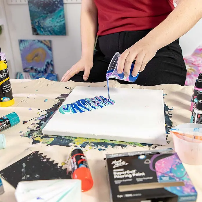 Mont Marte Acrylfarben-Set SuperCell® Tropical Ocean - mehrfarbig, 4 Stk. x 60 ml, Flasche - Person gießt Marmorierfarbe auf Leinwand und erschafft abstrakte Fluid Art. Farben und Künstlerbedarf liegen auf dem Tisch.