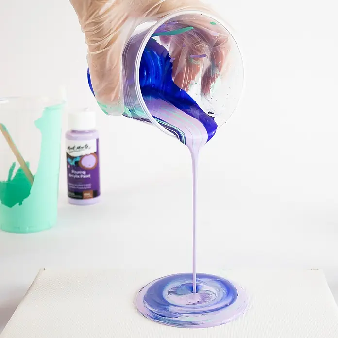Mont Marte Acrylfarben-Set Premium Ethereal - mehrfarbig, 4 Stk. x 60 ml, Flasche - Eine behandschuhte Hand gießt blaue, lila und hellgrüne Acrylfarbe aus einem Becher auf eine weiße Leinwand, wodurch ein wirbelndes Muster entsteht.
