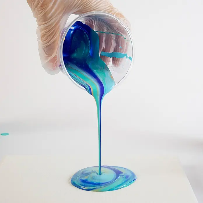 Mont Marte Acrylfarben-Set Premium Marina - mehrfarbig, 4 Stk. x 120 ml, Flasche - Blau-türkise Acrylfarbe mit Goldakzenten wird aus einem Becher auf eine weiße Leinwand gegossen und bildet einen Wirbel.