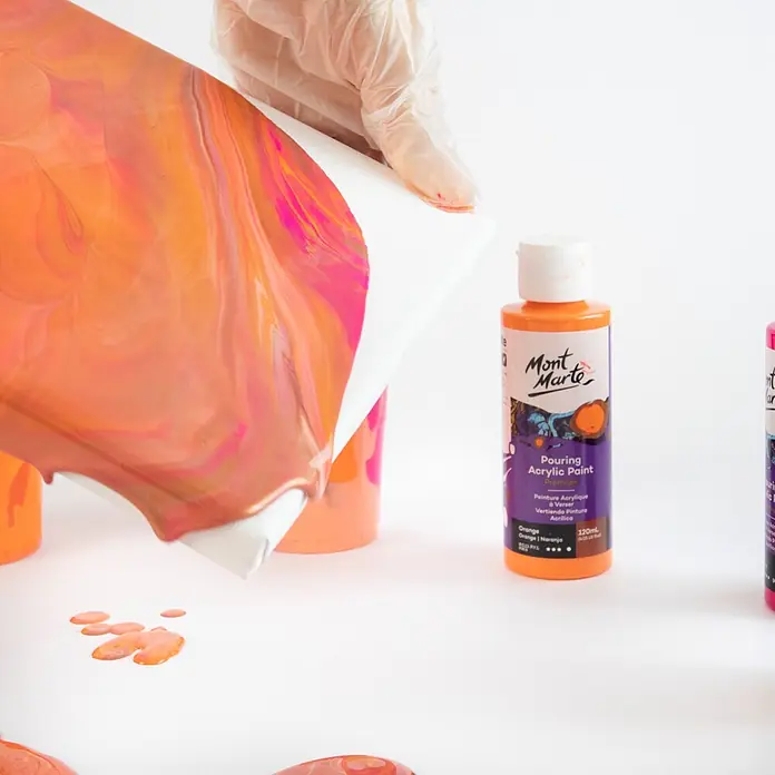 Mont Marte Acrylfarben-Set Premium Coral - mehrfarbig, 4 Stk. x 120 ml, Flasche - Behandschuhte Hand gießt orangefarbene und pinkfarbene Mont Marte Acrylfarbe auf Leinwand. Flaschen mit Gießfarbe sichtbar.