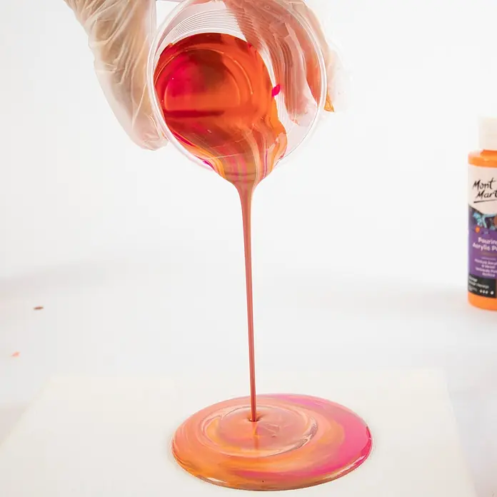 Mont Marte Acrylfarben-Set Premium Coral - mehrfarbig, 4 Stk. x 120 ml, Flasche - Eine behandschuhte Hand gießt leuchtende orange, pinke und goldene Acrylfarbe aus einem Becher auf eine Leinwand, die eine wirbelnde Pfütze bildet.