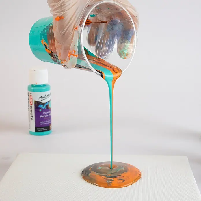Mont Marte Acrylfarben-Set Premium Galaxy - mehrfarbig, 4 Stk. x 60 ml, Flasche - Behandschuhte Hand gießt türkisblaue, orangefarbene und schwarze Farbe aus einem Becher auf eine weiße Leinwand.