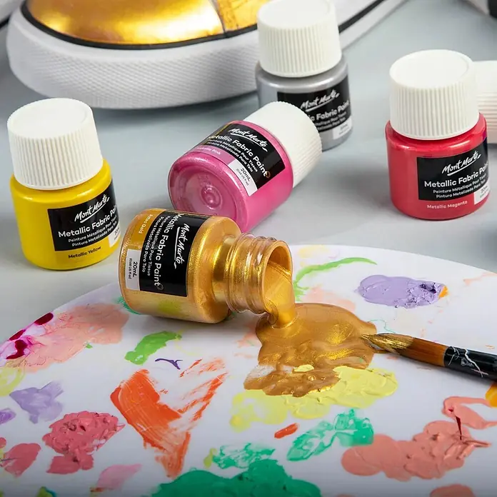 Mont Marte Textilmalfarben-Set Metallic Signature - 8x20ml - mehrfarbig, 8 Stk. x 20 ml - Mont Marte Metallische Stofffarben in verschiedenen Farben, eine goldene Flasche auf einer Palette verschüttet, daneben ein Pinsel. Goldener Schuh im Hintergrund.