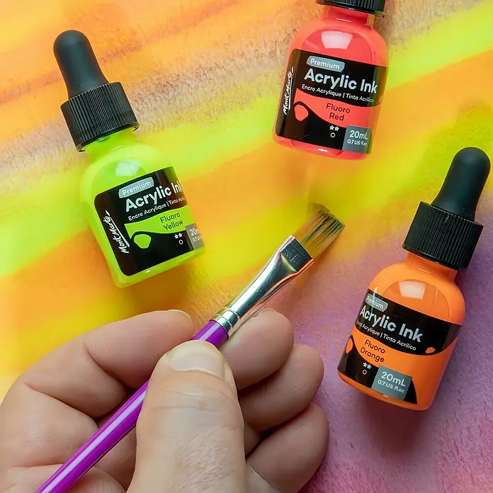 Mont Marte Zeichentuschen-Set Premium Neon - mehrfarbig, 12 Stk. x 20 ml - Hand hält Pinsel, umgeben von drei Mont Marte Fluoro Acryl-Tintenflaschen (gelb, rot, orange) auf einer leuchtenden bemalten Fläche.