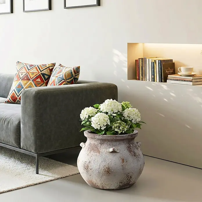 Hometopia Pflanztopf HTPL0358 - 1 Stk., Außenmaß (Ø x H): 37 x 30,5 cm, Verbundwerkstoff, Altweiß - Graues Sofa mit gemusterten Kissen, rustikalem Topf mit weißen Hortensien und Wandregal mit Büchern und Tasse im Wohnzimmer.