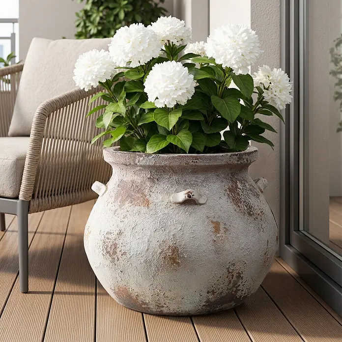 Hometopia Pflanztopf HTPL0358 - 1 Stk., Außenmaß (Ø x H): 37 x 30,5 cm, Verbundwerkstoff, Altweiß - Ein großer, verwitterter Pflanzkübel mit weißen Hortensien auf einem Holzdeck neben einem Gartenstuhl.