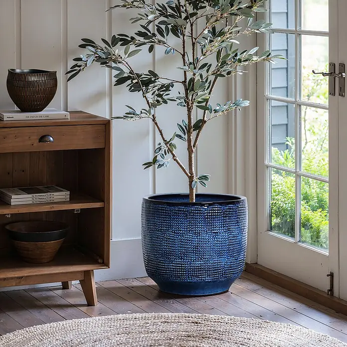 Hometopia Pflanztopf HTPL0036-L - 1 Stk., Außenmaß (Ø x H): 40 x 28,5 cm, Keramik, Marineblau - Großer, strukturierter blauer Glasurtopf mit künstlichem Olivenbaum auf Holzfußboden neben einem Schrank, vor einer Glastür.