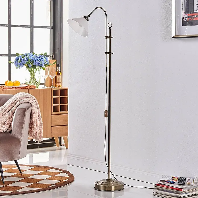 Lindby Stehleuchte Otis - bronze / altmessing, E27 - Stehleuchte im Vintage-Stil mit Messing-/Bronze-Finish und weißem Glasschirm, mit verstellbarem Arm, in einem Wohnzimmer gezeigt.
