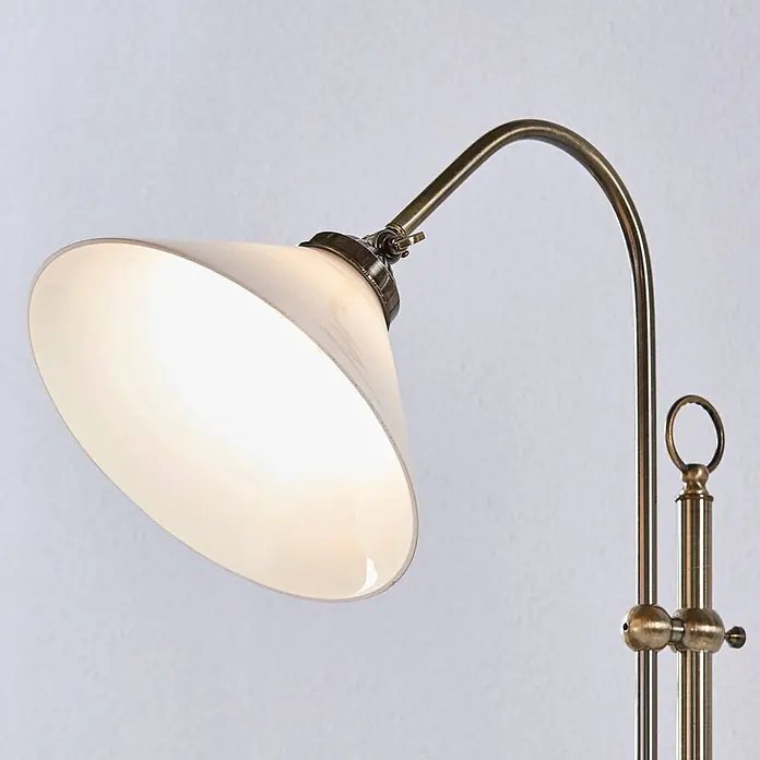 Lindby Stehleuchte Otis - bronze / altmessing, E27 - Detailansicht einer antiken Messing-Stehlampe mit gebogenem Arm und beleuchtetem, konischem Schirm aus weißem Opalglas.