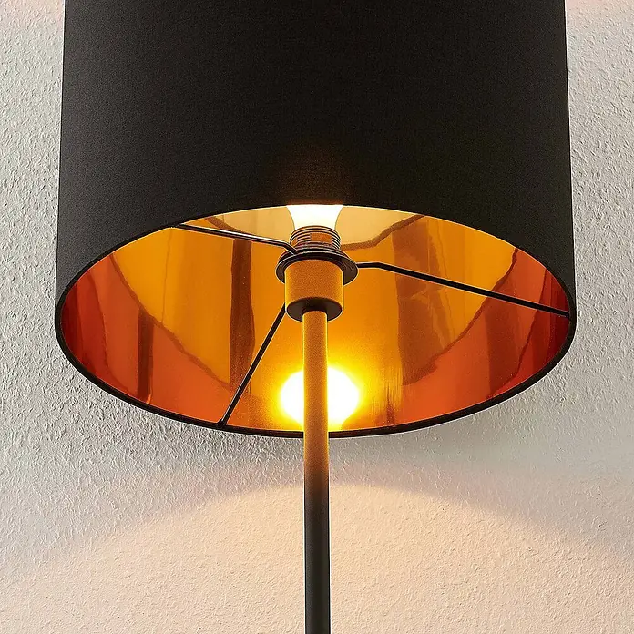 Lindby Stehlampe Wohnzimmer in schwarz, gold, Stehleuchte -  - Moderne Lampe mit schwarzem Schirm und goldener Innenseite, die warmes Licht auf eine strukturierte Wand wirft.
