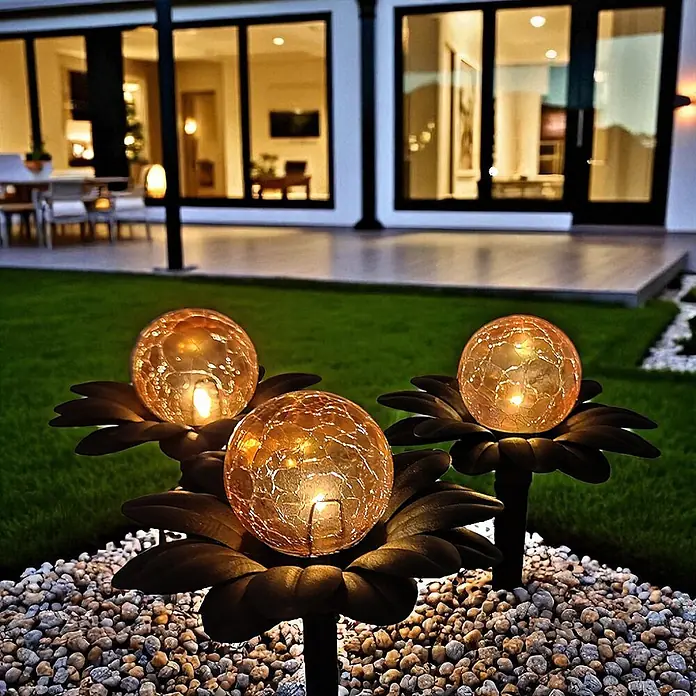DAPO Solar-Dekoleuchte Flora mit Kugel in Bernsteinoptik - L x B x H: 15 x 15 x 20 cm, Rostfarbig - Drei solarbetriebene Gartenleuchten mit rissigen Glaskugeln und dunklen Blumenbasen beleuchten einen Kiesweg im Garten bei Nacht.