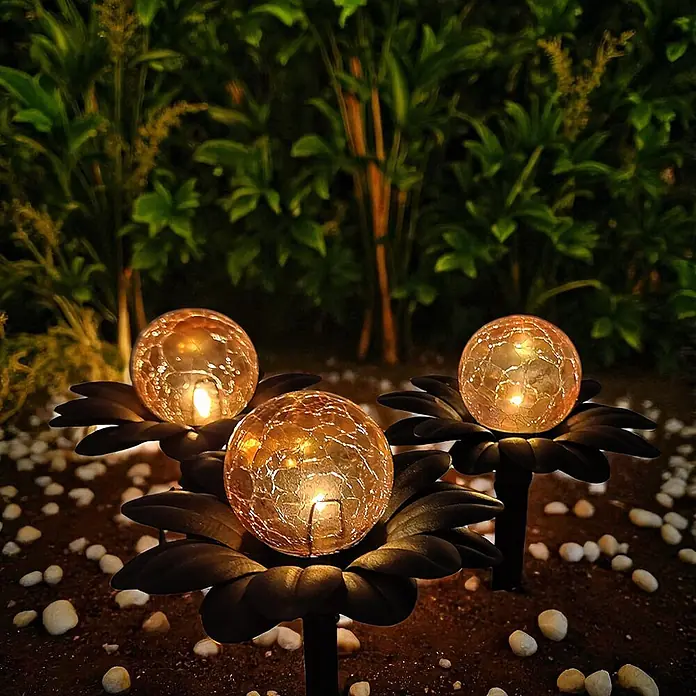 DAPO Solar-Dekoleuchte Flora mit Kugel in Bernsteinoptik - L x B x H: 15 x 15 x 20 cm, Rostfarbig - Drei Solar-Gartenleuchten mit Rissglas-Kugeln leuchten warm auf dunklen Blütenbasen und erhellen einen Gartenweg bei Nacht.