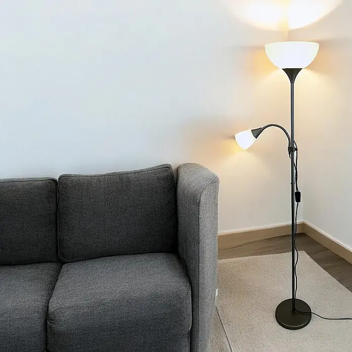 DAPO LED-Deckenfluter Stehleuchte Kora titan mit Leseleuchte - 23 W, Höhe: 178 cm, Titan, Weiß - Schwarze Metall-Stehlampe mit einer nach oben gerichteten Schale und einer flexiblen Leselampe, die ein dunkelgraues Sofa beleuchtet.