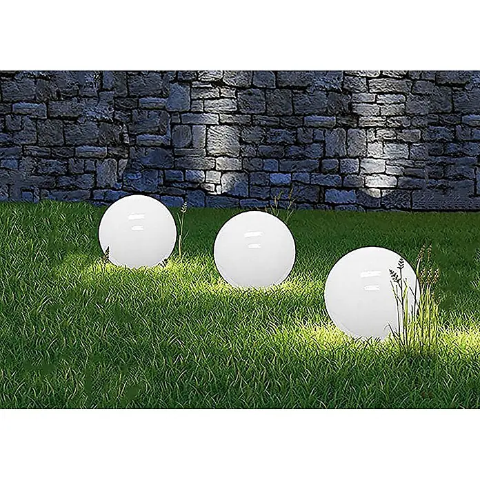 DAPO Solarleuchten-Set Ola 20 - L x B x H: 20 x 20 x 35 cm, Weiß, IP44 - Drei leuchtende weiße kugelförmige Außenleuchten stehen auf grünem Rasen vor einer Steinmauer.