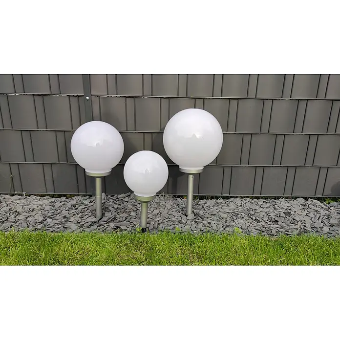 DAPO Solarleuchten-Set Ola 20 - L x B x H: 20 x 20 x 35 cm, Weiß, IP44 - Drei weiße kugelförmige Außenleuchten mit silbernen Sockeln, in einem Gartenbeet auf dunklen Schieferchips.